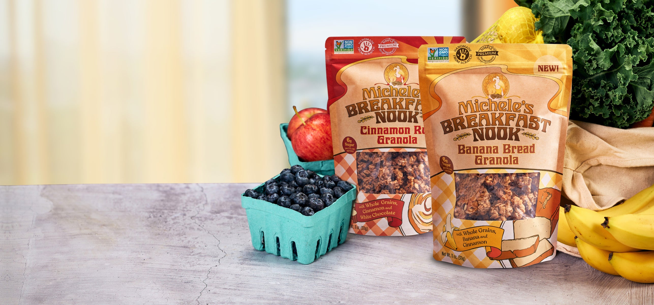 Two bags of Michele's Breakfast Nook granola are displayed on a light gray countertop with fresh fruits and vegetables, including blueberries in a turquoise container, an apple, and bananas, against a softly blurred kitchen background.