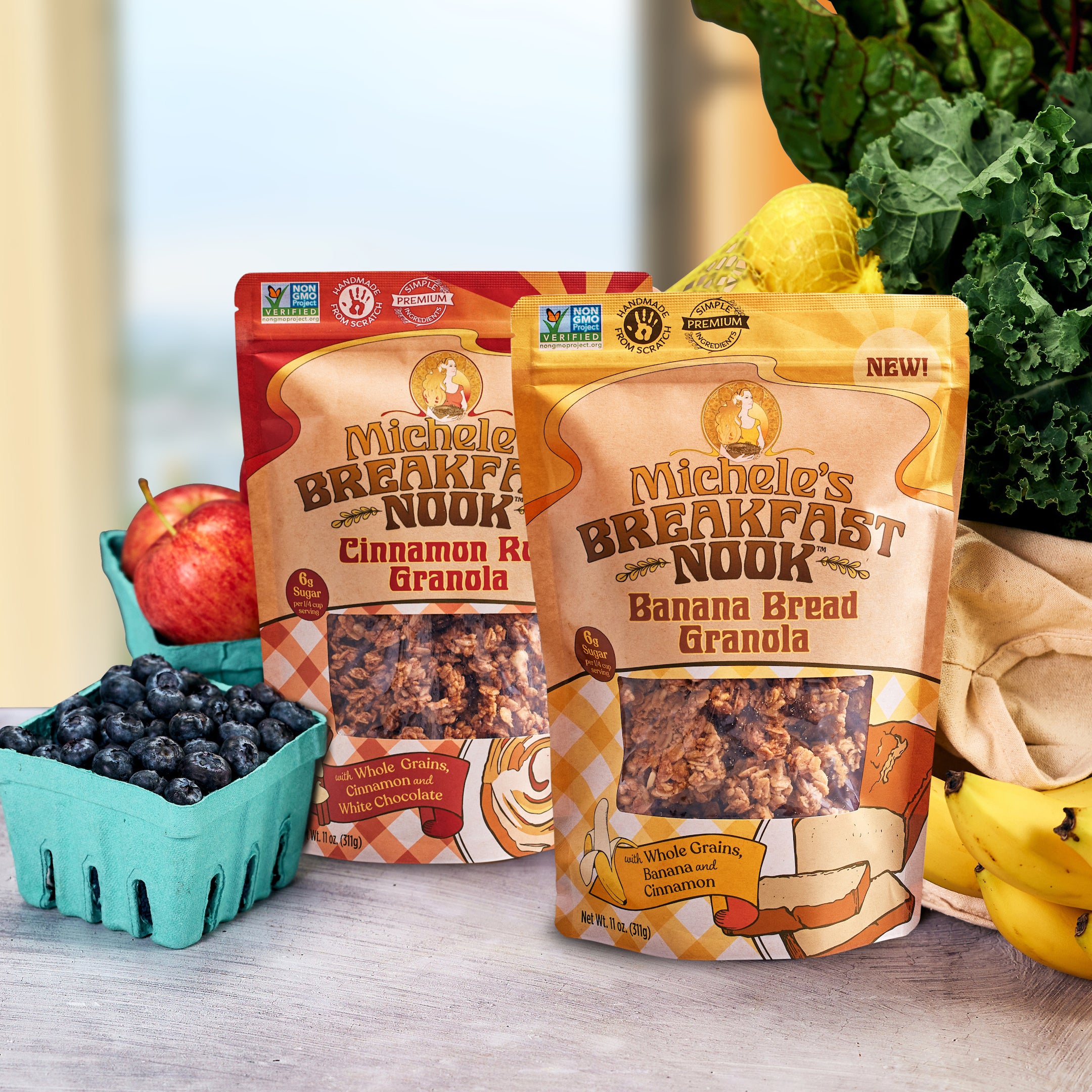 Two stand-up pouches of Michele's Breakfast Nook granola on a tabletop, surrounded by fresh ingredients including blueberries, an apple, bananas, and leafy greens. The front pouch is Banana Bread Granola and the rear pouch is Cinnamon Roll Granola.