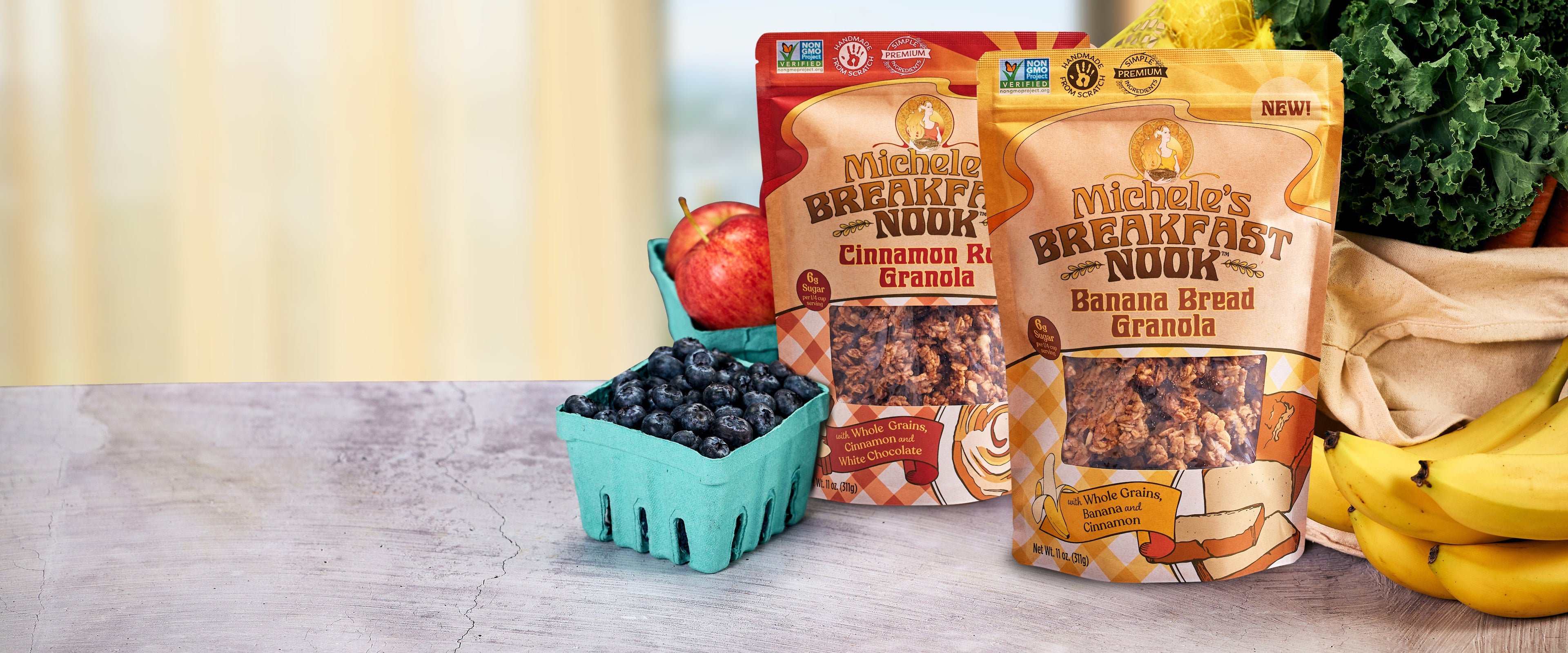 Two stand-up pouches of Michele's Breakfast Nook granola are displayed on a countertop with food props including blueberries, an apple, bananas, and kale. The front pouch is Banana Bread Granola and the back pouch is Cinnamon Roll Granola.