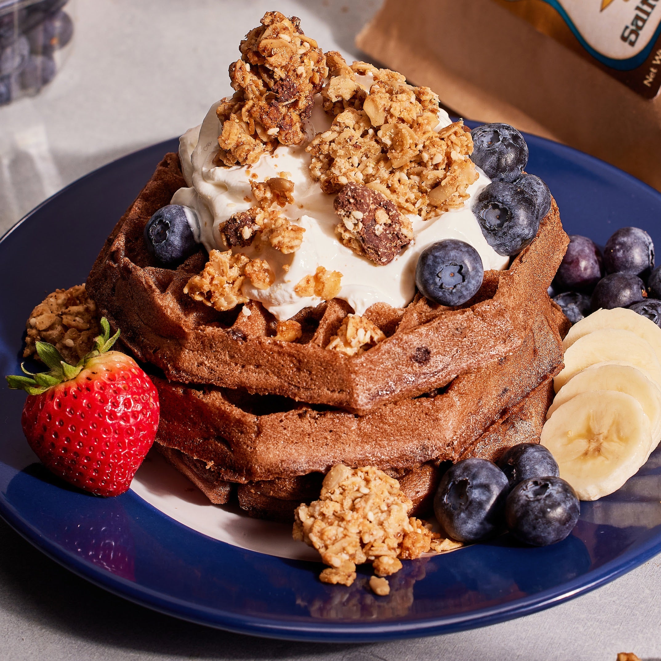 Chocolate waffles with whipped cream, Salted Chocolate Tahini Granola, blueberries, and a strawberry on a blue plate.
